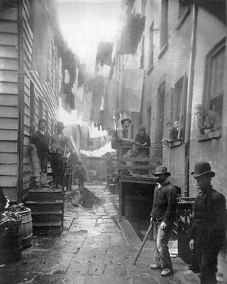 Men in bowler hats stand in the alleyway of a tenement house. Laundry hangs on lines above them. Men in bowler hats stand in the alleyway of a tenement house. Laundry hangs on lines above them.